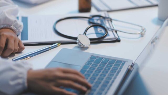 A professional and focused Asian female doctor in scrubs is working and reading medical research on her laptop in her office at a hospital.